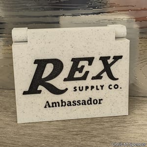 Puede incluir: Un letrero rectangular blanco moteado con las palabras "REX SUPPLY CO." y "Ambassador" en negrita negra. El letrero tiene una parte superior con bisagras y está sobre un fondo texturizado.