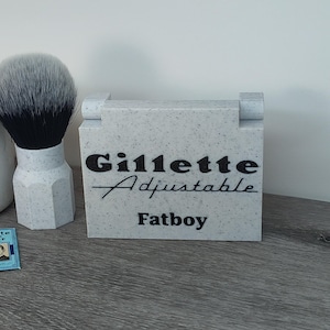 May include: A light gray block with the words "Gillette Adjustable Fatboy" in black script. A shaving brush with a white handle and black bristles sits to the left. Two blue Gillette Blue Blade packs are in the foreground.