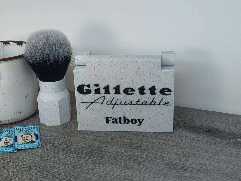 May include: A light gray block with the words "Gillette Adjustable Fatboy" in black script. A shaving brush with a white handle and black bristles sits to the left. Two blue Gillette Blue Blade packs are in the foreground.