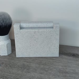 May include: A speckled gray shaving set featuring a brush with a black and white bristle head, a rectangular razor holder, and a vintage Gillette Blue Blade. The set is on a wooden surface.