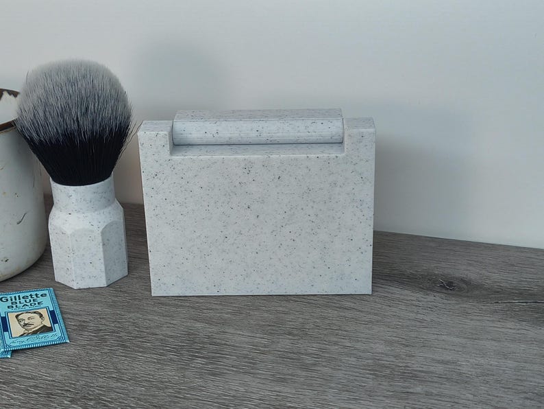May include: A speckled gray shaving set featuring a brush with a black and white bristle head, a rectangular razor holder, and a vintage Gillette Blue Blade. The set is on a wooden surface.