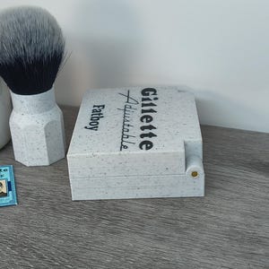 May include: A shaving set featuring a Gillette Fatboy Adjustable razor case, a shaving brush with a speckled handle and a two-tone brush head, and two Gillette Blue Blade packs. The case has the Gillette logo.