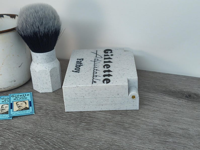 May include: A shaving set featuring a Gillette Fatboy Adjustable razor case, a shaving brush with a speckled handle and a two-tone brush head, and two Gillette Blue Blade packs. The case has the Gillette logo.