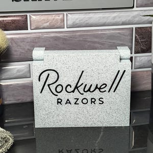 May include: A rectangular, light gray speckled stand featuring the black text Rockwell RAZORS printed on the front. The object has a smooth, stone-like texture with two small hinges visible at the top corners, designed for displaying shaving equipment.