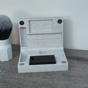 May include: A speckled gray shaving kit with a hinged lid, open to reveal a black object. A shaving brush with a white handle and a black bristle head is next to the kit. Two blue Gillette Blue Blade packages are also visible.