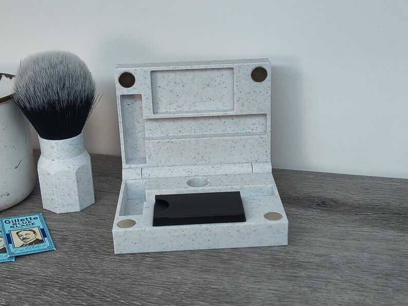 May include: A speckled gray shaving kit with a hinged lid, open to reveal a black object. A shaving brush with a white handle and a black bristle head is next to the kit. Two blue Gillette Blue Blade packages are also visible.
