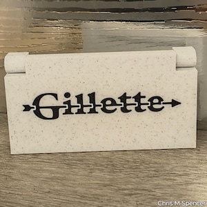 May include: A white speckled box with the word "Gillette" in black, with arrows on either side. The box has a hinged lid and is on a wooden surface. The image is well-lit and the text is clear.