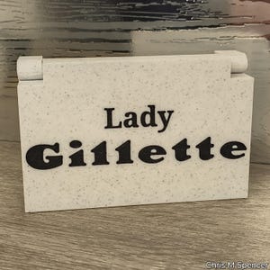 May include: A rectangular, speckled, off-white box with the words "Lady Gillette" in bold, black letters. The box has a hinged lid and is likely a container for a product.