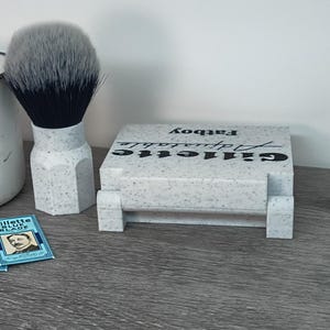 May include: A shaving set featuring a shaving brush with a gray and black bristle head, a speckled white and gray Gillette Adjustable Fatboy razor holder, and two Gillette Blue Blade packs. The items are arranged on a gray surface.