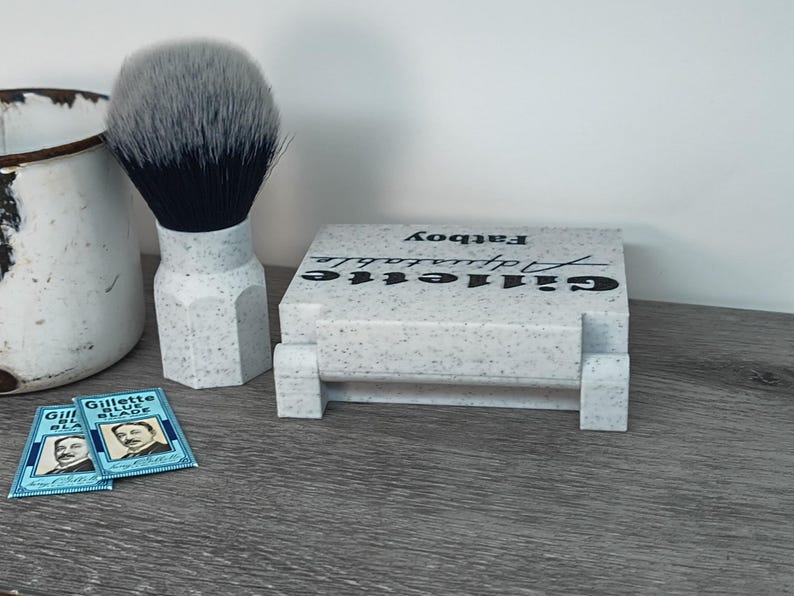 May include: A shaving set featuring a shaving brush with a gray and black bristle head, a speckled white and gray Gillette Adjustable Fatboy razor holder, and two Gillette Blue Blade packs. The items are arranged on a gray surface.