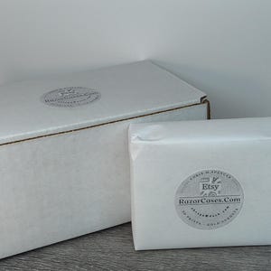 May include: Two white cardboard boxes, one large and one small, sit on a gray surface. The larger box is closed, while the smaller one is wrapped in white paper. Both have circular stickers with text.