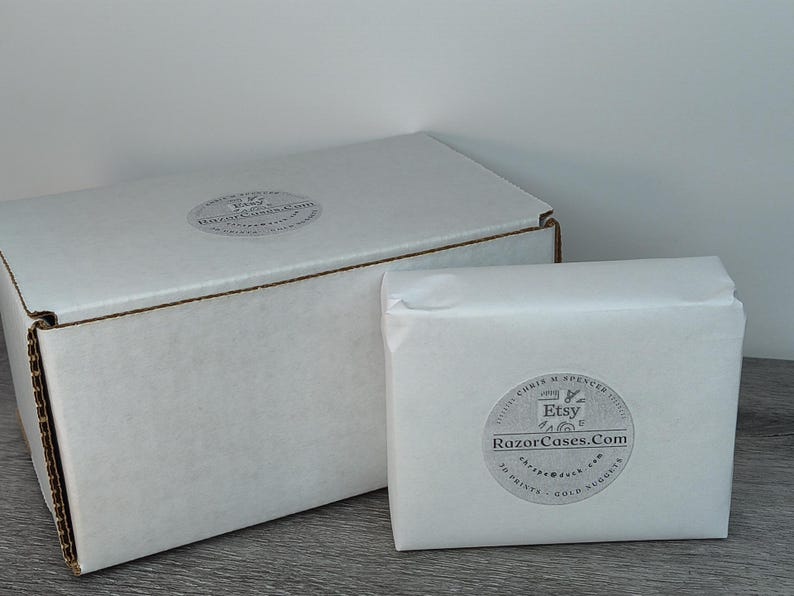 May include: Two white cardboard boxes, one large and one small, sit on a gray surface. The larger box is closed, while the smaller one is wrapped in white paper. Both have circular stickers with text.