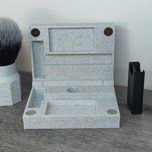May include: A light gray speckled shaving kit with a matching shaving brush and a black razor holder. The kit is open, revealing compartments for grooming essentials. A blue and white package of razor blades is also visible.