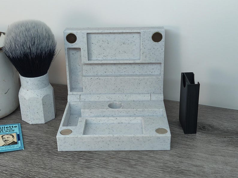 May include: A light gray speckled shaving kit with a matching shaving brush and a black razor holder. The kit is open, revealing compartments for grooming essentials. A blue and white package of razor blades is also visible.