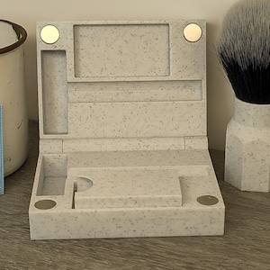 May include: A speckled, light-colored shaving kit with a hinged lid, compartments, and magnetic closures. A shaving brush with a white and black bristle head and a white handle is next to the kit. Vintage Gillette Blue Blade packs are visible.