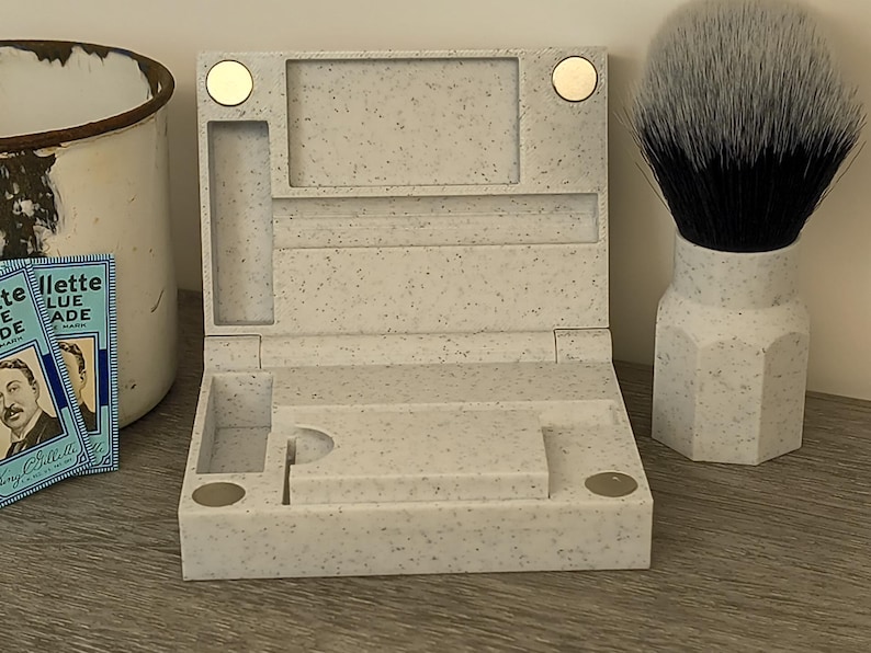 May include: A speckled, light-colored shaving kit with a hinged lid, compartments, and magnetic closures. A shaving brush with a white and black bristle head and a white handle is next to the kit. Vintage Gillette Blue Blade packs are visible.