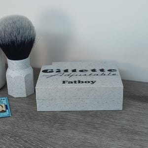 May include: A shaving set featuring a shaving brush with a black and gray bristle head and a speckled white handle. A box with the text "Gillette Adjustable Fatboy" is also present, along with a few blue Gillette blade packs.