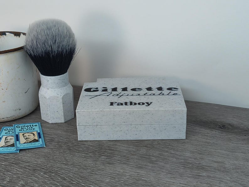 May include: A shaving set featuring a shaving brush with a black and gray bristle head and a speckled white handle. A box with the text "Gillette Adjustable Fatboy" is also present, along with a few blue Gillette blade packs.