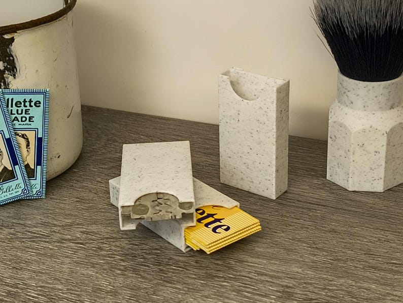 May include: A set of speckled gray razor blade holders, one open with yellow Gillette blades visible. A shaving brush and vintage Gillette Blue Blade box are also present, suggesting a grooming theme.