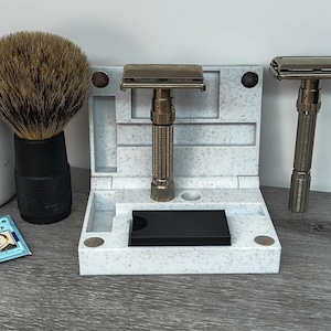 Op de afbeelding: Een scheerset met een scheerkwast, twee veiligheidsscheermessen en een gespikkelde wit en grijze scheermesstandaard. Twee Gillette Blue Blade-verpakkingen zijn ook zichtbaar. De scheermessen en borstel hebben donkere metalen en zwarte accenten.