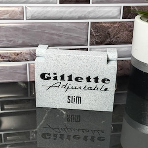 May include: A light gray Gillette Adjustable Slim razor case. The case has the brand name and product name printed in black. The case is rectangular with a hinged top. The background is a tiled wall.