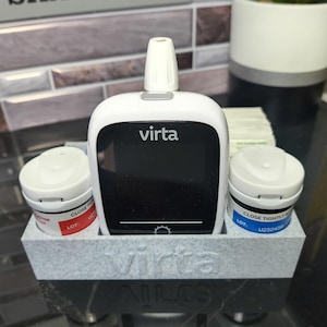 May include: A white Virta blood glucose monitor with a black screen, flanked by two white containers in a gray holder. The containers have red and blue labels. The word "Virta" is printed on the monitor and holder.