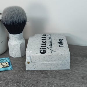 May include: A shaving set featuring a speckled gray and white Gillette Adjustable Fatboy razor case, a matching shaving brush with a gray and black brush head, and two Gillette Blue Blade packs. The items are arranged on a gray surface.