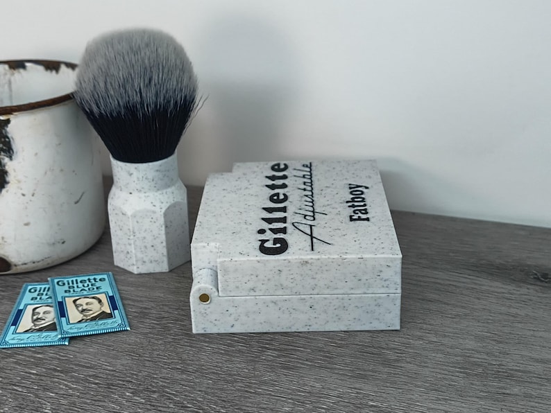 May include: A shaving set featuring a speckled gray and white Gillette Adjustable Fatboy razor case, a matching shaving brush with a gray and black brush head, and two Gillette Blue Blade packs. The items are arranged on a gray surface.