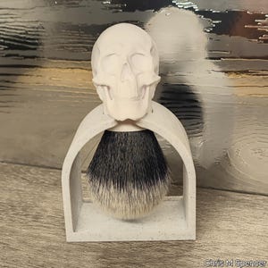 May include: A white skull-shaped shaving brush holder with a black and white brush. The holder has an arched design and a rectangular base. The brush bristles are a mix of black and white. A unique, gothic-inspired bathroom accessory.
