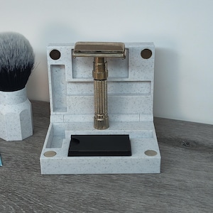 May include: A shaving set featuring a safety razor, shaving brush, and a stand. The razor has a gold-colored head and a textured handle. The stand is white with a speckled pattern and has compartments for the razor and other grooming items.
