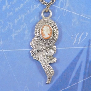 May include: A silver-toned metal pendant necklace featuring an oval cameo with a white profile silhouette on an orange background. The pendant has a textured, beaded border and a decorative, flowing ribbon-like design extending from the base.