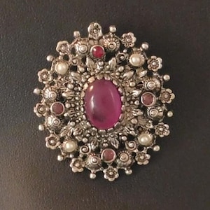 May include: Silver-toned brooch with an oval, magenta-colored center stone. The brooch features a detailed floral design with small red and white accents. A vintage-style jewelry piece.