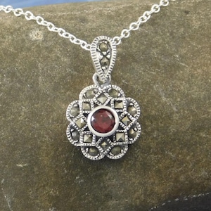 May include: A silver pendant necklace featuring a flower-shaped pendant with a central red gemstone. The pendant is adorned with small, sparkling accents and hangs from a delicate silver chain. The jewelry is displayed on a textured surface.