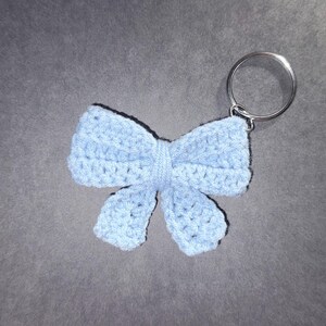 May include: A light blue crocheted bow keyring. The bow is made of soft yarn and has a detailed, textured appearance. A silver-coloured metal ring is attached to the top for easy attachment to keys or bags. The keyring is a handmade accessory.