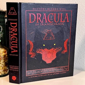May include: A hardcover book titled Dracula of Transylvania by Ricardo Delgado with a black cover featuring red text and a red graphic of a wolf-like creature. The spine displays the title in red text and a small red flower icon at the bottom.