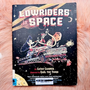 May include: A book cover for "Lowriders in Space" by Cathy Camper, illustrated by Raúl The Third. The cover features a black background with a space scene and a lowrider car with cartoon characters. The title is in red and white.