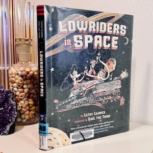 May include: A hardcover book titled "Lowriders in Space" with a dark blue cover featuring a car in space with cartoon characters. The spine reads "Lowriders in Space" and the author is Cathy Camper.