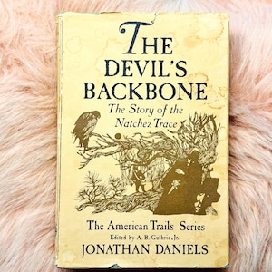 Könnte beinhalten: Ein gebundenes Buch mit dem Titel The Devil's Backbone: The Story of the Natchez Trace von Jonathan Daniels. Der cremefarbene Einband zeigt eine dunkelbraune Tuschezeichnung eines Vogels, Bäumen und einer Figur sowie den Text The American Trails Series.