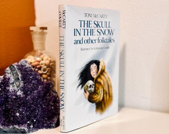 Op de afbeelding: Een wit hardback boek getiteld "The Skull in the Snow and other folktales" van Toni McCarty. De omslag van het boek toont een illustratie van een vrouw in een bontjas die een schedel vasthoudt.