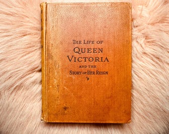 Könnte beinhalten: Ein antikes Buch mit dem Titel "The Life of Queen Victoria and the Story of Her Reign" mit einem abgenutzten, orangefarbenen Einband. Das Buch liegt vor einem Hintergrund aus weichem, pfirsichfarbenem Fell.