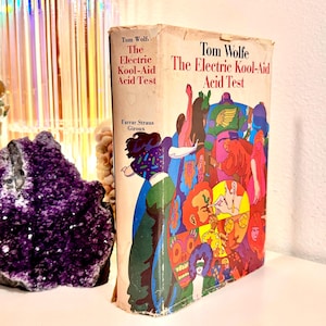 May include: A copy of Tom Wolfe's "The Electric Kool-Aid Acid Test" book with a vibrant cover illustration. The book is displayed alongside a purple amethyst geode and other decorative items, creating a colourful display.