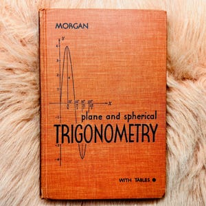 May include: An orange hardcover book titled "plane and spherical TRIGONOMETRY" with a graph illustration. The book's cover also includes the name "MORGAN" and the words "WITH TABLES". The book is resting on a fluffy, light-colored surface.