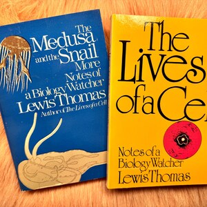 May include: Two books with colorful covers. The blue book is titled "The Medusa and the Snail" by Lewis Thomas. The yellow book is titled "The Lives of a Cell" by Lewis Thomas. Both books are on a fuzzy, peach-colored surface.
