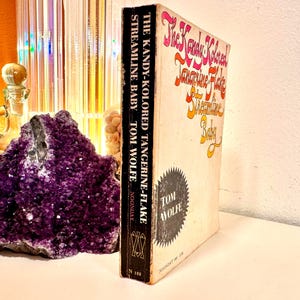 May include: A vintage copy of "The Kandy-Kolored Tangerine-Flake Streamline Baby" by Tom Wolfe. The book has a cream-colored cover with orange and pink lettering. The spine is black with white text. A large purple amethyst geode is in the background.