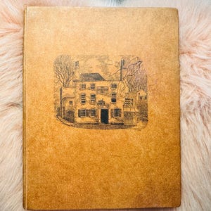 May include: A vintage book with a tan cover featuring a detailed black and white illustration of a building with multiple windows and a central door. The book is resting on a fluffy, light pink surface.