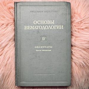 Puede incluir: Un libro vintage gris verdoso titulado "ОСНОВЫ НЕМАТОДОЛОГИИ" (Fundamentos de Nematodología) con letras blancas. El libro está sobre una superficie rosa claro y esponjosa. La portada presenta texto y un pequeño emblema.
