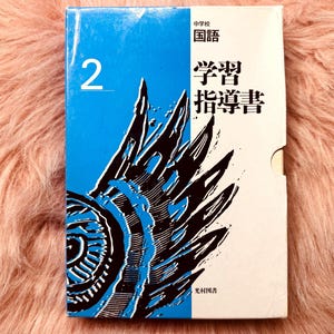 May include: A Japanese textbook with a blue and white cover. The cover features the number "2" and Japanese text. A blue and black graphic design is on the cover. The book is on a pink, fuzzy surface.