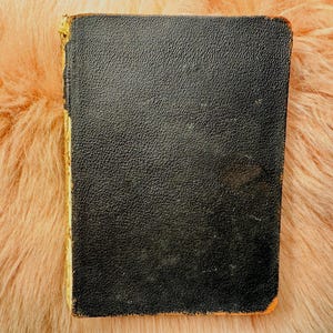 Puede incluir: Un libro pequeño y negro vintage con una cubierta texturizada. Los bordes del libro están amarillentos y las esquinas muestran desgaste. El libro está sobre un fondo de material esponjoso color melocotón.