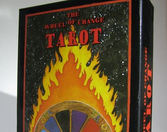 May include: The Wheel of Change Tarot deck box features a black background with a colorful wheel of fortune surrounded by flames. The text "The Wheel of Change Tarot" is written in red and white on the front of the box.