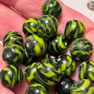 May include: A collection of round beads with a green and yellow striped pattern. The beads are small, with a hole through the center for stringing. The beads are held in a hand.